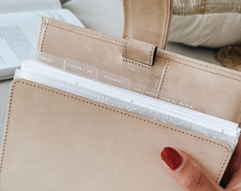 May include: A beige leather personal planner with a snap closure and metal ring binding. The interior features translucent dividers labeled with text including INBOX, VISION / 23, AGENDA, L+P / ODSP, and SOCIAL MEDIA, holding white paper pages.
