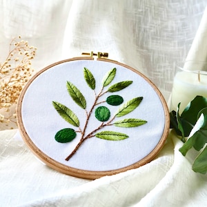 May include: A circular wooden embroidery hoop containing white fabric with a hand-stitched design of an olive branch. The branch features brown stems, multiple pointed green leaves in varying shades, and three round, dark green olives.