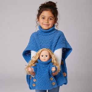 Puede incluir: Una niña pequeña con un poncho azul tejido con botones de madera, sosteniendo una muñeca con un suéter azul tejido a juego. La muñeca tiene el pelo rubio y lleva un jersey de cuello alto azul con botones de madera. El fondo es gris claro liso.
