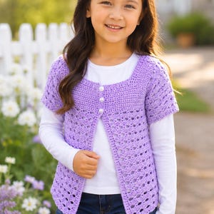 May include: A light purple, short-sleeved crochet cardigan with three white buttons. The open-weave design is worn over a white long-sleeved shirt and blue jeans. The cardigan is a handmade garment.