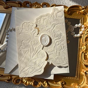 May include: Ivory-colored invitation with embossed floral design and a wax seal. The invitation is layered with an envelope and rests on a gold-framed mirror. A strand of pearls is draped nearby, suggesting a wedding or special event.
