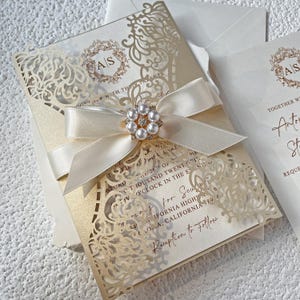 Pode incluir: Elegante convite de casamento marfim com detalhes intrincados cortados a laser. Apresenta uma fita de cetim creme adornada com um enfeite de pérolas e strass. Inclui texto impresso e envelope a condizer.