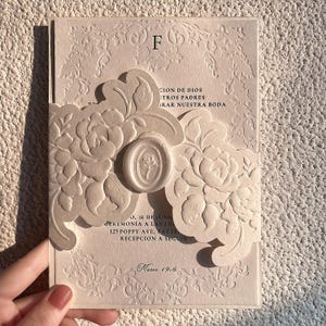 May include: A cream-coloured wedding invitation card featuring an embossed floral belly band secured with a central wax seal. The card displays black printed text including the letter F, wedding details, and the scripture reference Mateo 19:6.