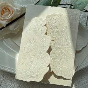 May include: A cream-coloured invitation card featuring intricate embossed floral patterns on the front panels. The card has scalloped edges and a textured surface, showcasing detailed rose and tulip designs in a monochromatic finish.