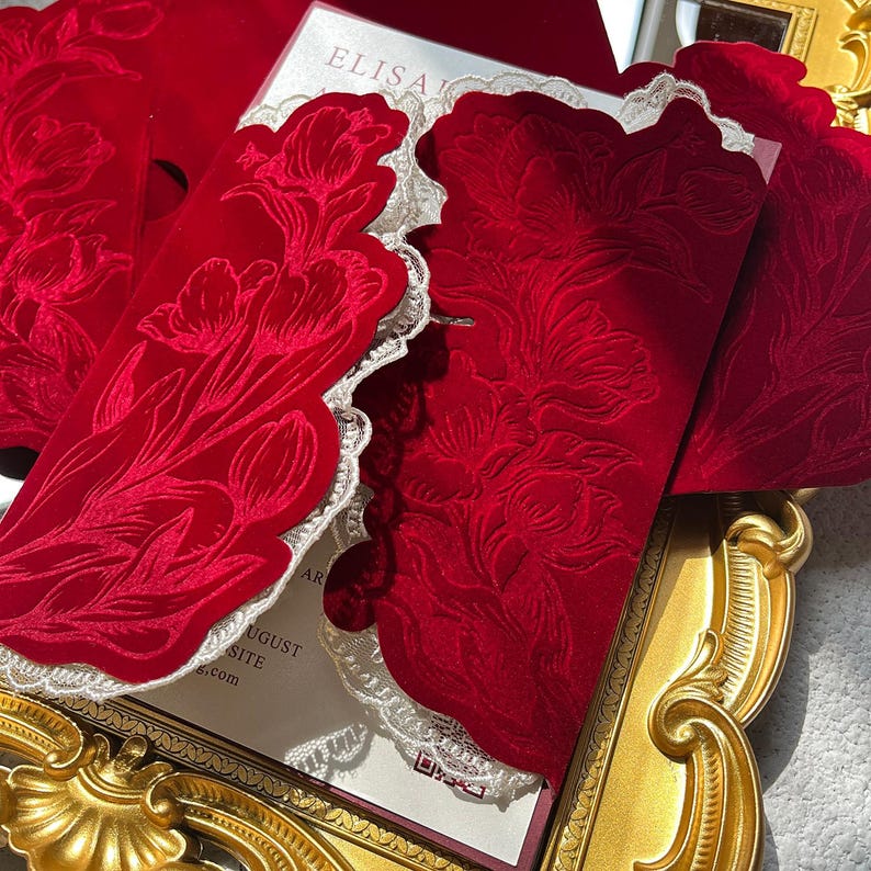 May include: Red velvet invitation with floral embossing and a scalloped lace edge. The invitation features the text "ELISA" and other details. The invitation is displayed on a gold ornate frame.