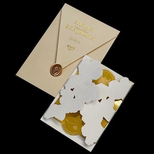 May include: An ivory wedding invitation suite with gold foil text that reads "SARAH BENJAMIN" and the date "28.09.26". The envelope is sealed with a wax stamp. The invitation features white floral cutouts.
