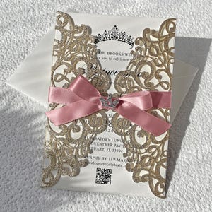 Peut inclure: Une invitation avec un motif de filigrane doré scintillant, un ruban de satin rose et une couronne en strass. L'invitation comprend le texte "Mr. Brooks Weiser vous invite à célébrer..." et les détails de l'événement.