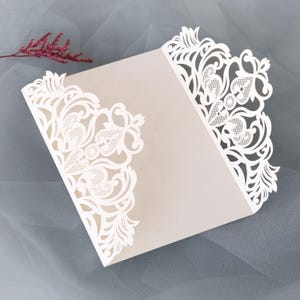 Peut inclure: Une invitation de mariage beige avec des motifs floraux blancs découpés au laser sur les bords. L'invitation est ouverte, révélant l'intérieur. Une brindille de fleurs roses est posée près du haut. L'ensemble est élégant et romantique.