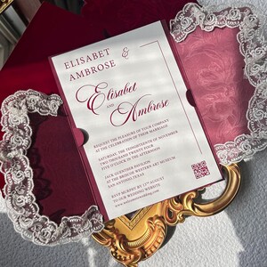 May include: A burgundy wedding invitation with the names "Elisabet & Ambrose" in elegant script. The invitation includes event details and is framed by a decorative white lace border. The invitation is on a gold ornate tray.
