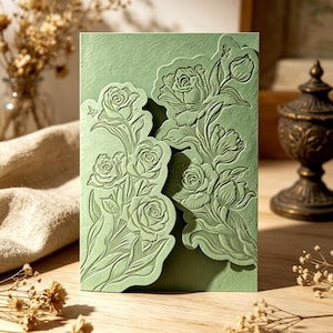 Op de afbeelding: Een saliegroene wenskaart met reliëf bloemmotieven. De kaart heeft gedetailleerde reliëfsnijwerken van rozen en bladeren. Het ontwerp heeft een botanisch thema, met een getextureerde papieren afwerking. De kaart is rechthoekig en staat rechtop.