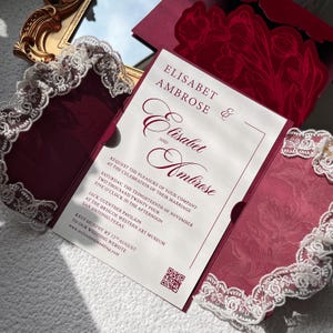 May include: Burgundy wedding invitation with white lace trim and floral accents. The invitation includes the names "Elisabet & Ambrose" and event details. A QR code and website address are also visible.