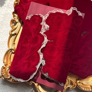 May include: Close-up of red velvet invitations with floral designs and white lace trim. The invitations are arranged on a gold-colored decorative tray, creating an elegant and luxurious aesthetic.