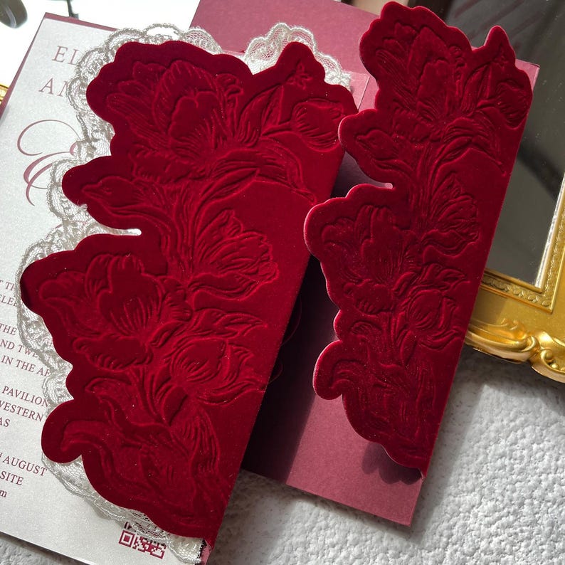 May include: Close-up of a red velvet invitation with a floral design. The invitation has a cut-out edge and is paired with a white lace trim. The text "ELA AN" is visible on the invitation.