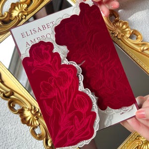 May include: Red velvet invitation with a floral design and white lace trim. The invitation features the text "ELISABET AMBRO". The invitation is displayed on a gold framed mirror.