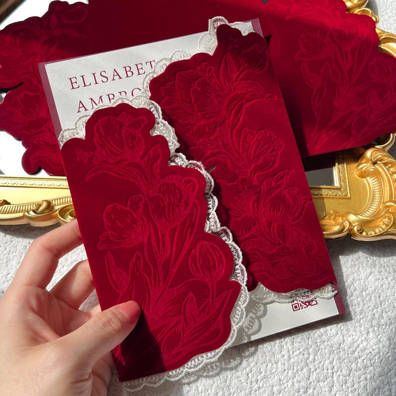 May include: Red velvet invitation with floral embossing and a white lace trim. The invitation features the text "ELISABET AMBRO". The design includes a matching envelope and additional cards, suggesting a wedding or special event theme.