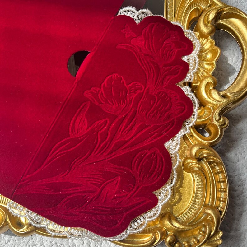 May include: A red velvet envelope with a floral design and scalloped edges, accented with white lace trim. The envelope is partially open, revealing a glimpse of its interior. The envelope rests on a gold ornate frame.