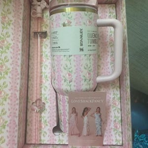 May include: A floral-patterned Stanley Quencher tumbler in a gift box. The tumbler is pink with a white and pink floral design, a pink handle, and a clear lid. The box and included straw also feature the floral pattern. A small card with women in dresses is included.
