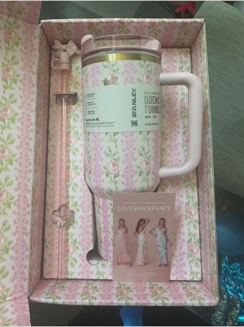 May include: A floral-patterned Stanley Quencher tumbler in a gift box. The tumbler is pink with a white and pink floral design, a pink handle, and a clear lid. The box and included straw also feature the floral pattern. A small card with women in dresses is included.