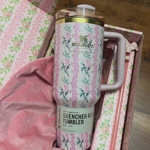 May include: A pink and white floral Stanley Quencher H2.0 tumbler with a handle, inside a matching floral box. The tumbler has the text "STANLEY LOVESHACKFANCY" and "QUENCHER H2.0 TUMBLER 40 OZ 1.18 L".