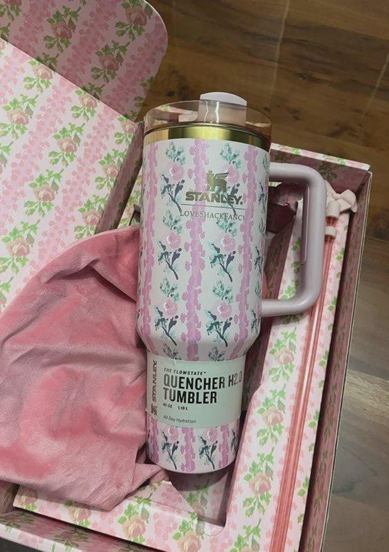 May include: A pink and white floral Stanley Quencher H2.0 tumbler with a handle, inside a matching floral box. The tumbler has the text "STANLEY LOVESHACKFANCY" and "QUENCHER H2.0 TUMBLER 40 OZ 1.18 L".