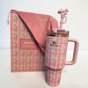 May include: A pink Stanley tumbler with a floral design, a handle, and a bow-shaped straw. The tumbler is next to a matching box and a pink velvet bag. The box has the text "Stanley LOVESHACKFANCY".