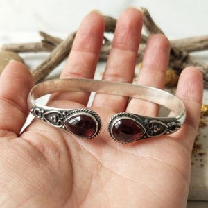 Peut inclure: Bracelet manchette en argent avec deux pierres précieuses rouges en forme de goutte d'eau. Le bracelet présente des détails ornés autour des pierres précieuses et sur les côtés. Le design ouvert permet de le porter facilement. Un bijou élégant.