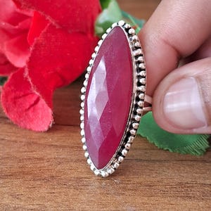 Può includere: Un anello a forma di marquise con una grande pietra preziosa rosso scuro sfaccettata, montata su una fascia in metallo color argento. L'incastonatura presenta un bordo testurizzato a perline che circonda la pietra centrale, creando un profilo allungato.