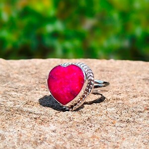 May include: A silver ring featuring a prominent heart-shaped, vibrant magenta-pink gemstone. The stone is set within a decorative, twisted rope-style metal border, and the band is crafted from a smooth, polished silver-toned metal finish.