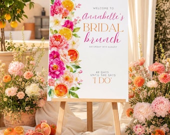 May include: A white rectangular sign on a wooden easel featuring a floral and citrus design on the left side. The text reads: WELCOME TO Annabelle's BRIDAL brunch SATURDAY 14TH AUGUST 48 DAYS UNTIL SHE SAYS 'I DO'. The flowers are pink and white.