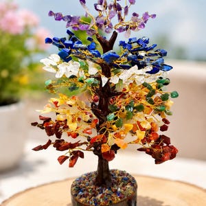 May include: A decorative tree sculpture featuring a dark brown trunk and branches made of gold-colored wire. The branches are adorned with multi-colored gemstone chips in purple, blue, white, green, yellow, and red. The base is cylindrical with the text "orgonite Aura Co" printed on the front.