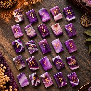 May include: A set of twenty-five rectangular amethyst stones arranged in a grid, each featuring a unique gold-colored engraved rune symbol. The stones display natural purple and white banding, with polished surfaces and rounded edges.