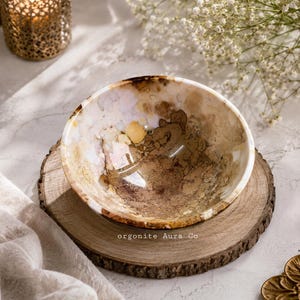 May include: A round bowl featuring a marbled pattern in shades of cream, tan, brown, and soft lavender. The interior surface has a glossy finish with organic, swirling colour variations. The text "orgonite Aura Co" is printed on the base.
