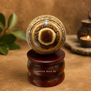 May include: A spherical stone with concentric bands of brown, tan, and cream colours, featuring a prominent circular centre. The sphere rests on a dark brown, cylindrical wooden stand with the text orgonite Aura Co printed on the base.