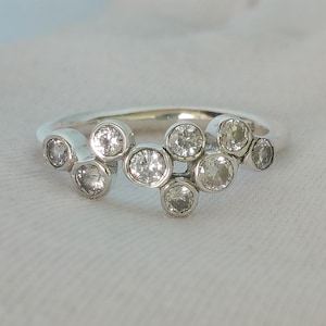 Può includere: Un anello color argento con un gruppo di otto pietre rotonde e trasparenti incastonate singolarmente. Le pietre sono disposte in modo asimmetrico e sfalsato sulla parte superiore della fascia, creando un aspetto organico a più livelli.