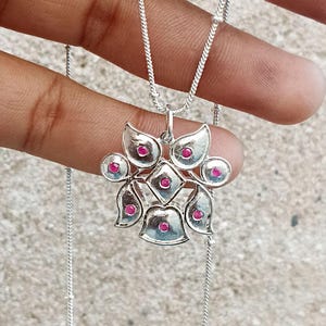 Può includere: Una collana in argento con un ciondolo dal design simmetrico ispirato a un fiore. Il ciondolo è decorato con sette piccole pietre rotonde di colore rosa vivace incastonate in petali d'argento lucido e una cornice centrale a rombo.