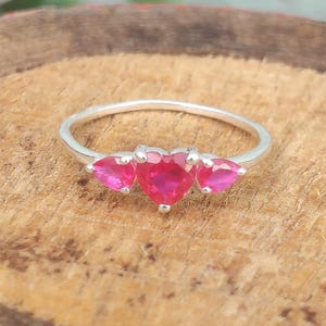 Op de afbeelding: Een zilveren ring met drie hartvormige, felroze stenen op een rij. De middelste hartvormige steen is groter dan de twee flankerende hartvormige stenen, allemaal vastgezet met metalen pootjes op een dunne, gepolijste zilveren band.