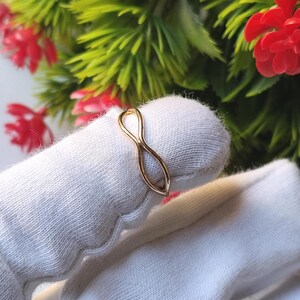 Op de afbeelding: Een dunne gouden ring met een delicaat oneindigheidssymbool-ontwerp. De band is gemaakt van glad, gepolijst goudkleurig metaal en vormt een doorlopende lus die in het midden smaller wordt om de klassieke acht-vorm van het oneindigheidssymbool te creëren.