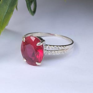 Puede incluir: Un anillo de plata con una gran piedra preciosa de color rojo intenso y corte ovalado, sujeta por cuatro garras. La banda está dividida en dos secciones a cada lado, cada una incrustada con pequeñas piedras claras y brillantes.