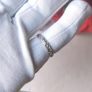 Peut inclure: Une bague en métal argenté présentant un motif de volutes gravé en continu sur l'anneau. La bague est présentée sur un doigt recouvert d'un gant en tissu blanc, mettant en valeur les détails complexes et répétitifs gravés en surface.