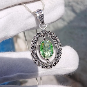 May include: A silver-toned pendant necklace featuring an oval-shaped, faceted light green gemstone set within an intricate, filigree-style oval frame. The pendant hangs from a thin silver-toned chain, showcasing detailed metalwork.