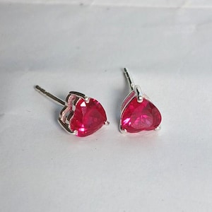 May include: A pair of stud earrings featuring faceted, heart-shaped red stones set in silver-toned metal. Each earring has a post back and a clear, vibrant red gemstone cut into a heart shape, held securely by a metallic prong setting.