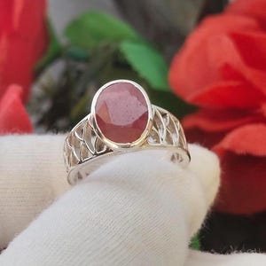 May include: A silver ring featuring an oval-shaped, reddish-pink gemstone set in a bezel. The band of the ring has an intricate, openwork lattice design that extends around the sides, providing a detailed metal structure beneath the central stone.