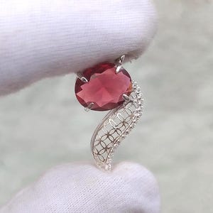 May include: A silver pendant featuring a large, oval-cut, deep red garnet gemstone held by four prongs. The pendant includes a curved, openwork metal filigree section with a repeating circular pattern and small, clear, round-cut accent stones.
