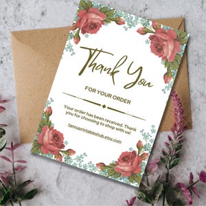 May include: A white card with a floral design and the words "Thank You" in a handwritten style. The card has red roses and blue accents. A brown envelope is behind the card.