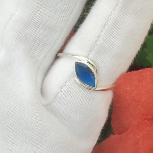 Puede incluir: Un anillo de plata con una piedra azul única de corte marquesa montada en una banda de estilo bypass. La banda de metal se curva alrededor de los extremos puntiagudos de la piedra, creando un diseño elegante que resalta su color azul.
