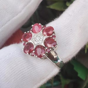 May include: A silver ring featuring a cluster of six oval-cut deep red rubies arranged in a floral pattern, with a central star-shaped setting containing small clear accent stones. The band is polished silver and has a smooth, reflective finish.