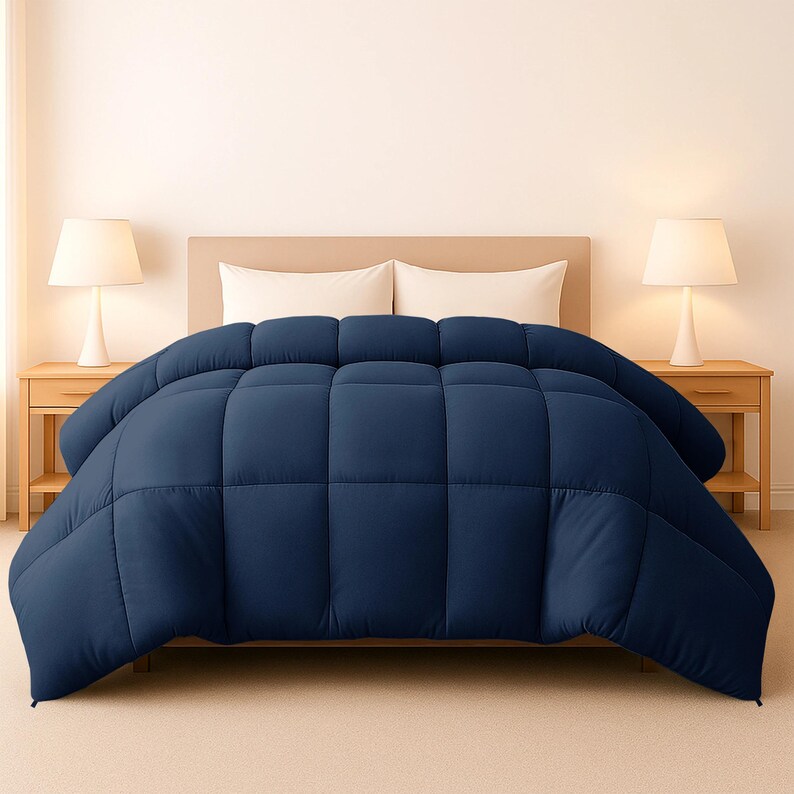 Op de afbeelding: Een marineblauwe dekbedovertrek op een bed. De dekbedovertrek is gewatteerd met een vierkant patroon. Twee witte kussens zijn zichtbaar achter de dekbedovertrek. Twee lampen staan op houten nachtkastjes aan weerszijden van het bed.