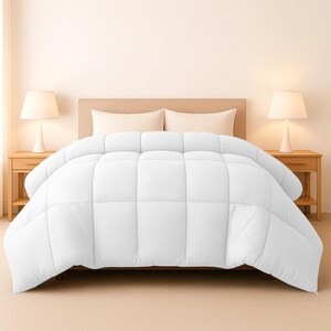 Op de afbeelding: Een witte, gewatteerde dekbedovertrek op een bed. De overtrek heeft een vierkant patroon en is netjes gerangschikt. Het bed staat in een slaapkamer, met lampen aan weerszijden. De algehele esthetiek is schoon en eenvoudig.