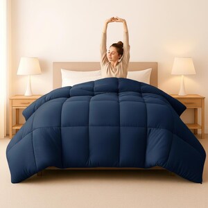 Op de afbeelding: Een marineblauwe dekbedovertrek op een bed. De dekbedovertrek heeft een gewatteerd ontwerp. Twee witte lampen staan op houten nachtkastjes. Een persoon rekt zich uit met de armen boven het hoofd.
