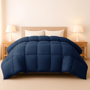 Op de afbeelding: Een marineblauwe dekbedovertrek op een bed. De dekbedovertrek is gewatteerd met een vierkant patroon. Twee witte kussens zijn zichtbaar achter de dekbedovertrek. Twee lampen staan op houten nachtkastjes aan weerszijden van het bed.